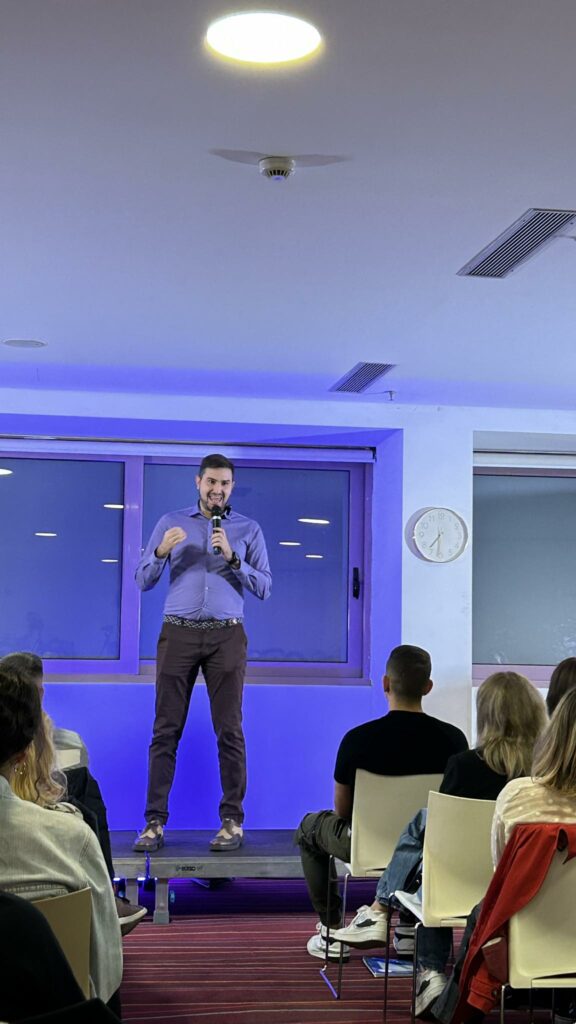 Guy speaker standing on a scene giving a speech. Purple lighting on the room.